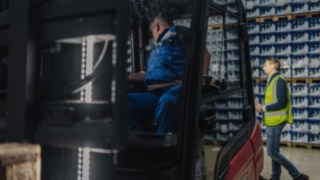 Un pedone con un gilet ad alta visibilità cammina per il magazzino dietro un carrello elevatore.