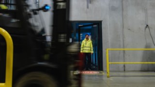 Un uomo in tenuta di sicurezza esce da una porta per entrare nel capannone di una fabbrica. Passa un carrello elevatore. Un uomo in tenuta di sicurezza esce da una porta per entrare nel capannone di una fabbrica. Passa un carrello elevatore.