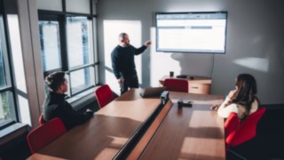 Tre persone tengono una riunione di gruppo sui progetti di intralogistica e automazione in una sala riunioni con una presentazione sullo schermo.