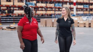 Due collaboratrici conversano in un moderno magazzino, vestite con l'abbigliamento aziendale Linde, simbolo della diversità culturale, della collegialità vissuta e della stretta collaborazione in un team globalmente interconnesso e localmente radicato.