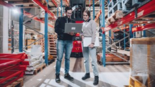 Due dipendenti sono in piedi davanti a un veicolo Linde in un magazzino automatizzato a scaffali alti e discutono di questioni tecniche sul laptop, simboleggiando il lavoro sulle moderne soluzioni di automazione in un ambiente innovativo e tecnologico.