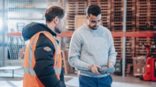 Due dipendenti impegnati in uno scambio tecnico in un ambiente industriale o di magazzino, uno dei quali indossa un giubbotto di sicurezza e l'altro utilizza un dispositivo di controllo, a simboleggiare progetti internazionali, responsabilità tecniche e opportunità di carriera a livello globale.