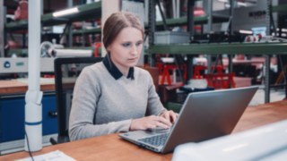 Una dipendente lavora concentrata al computer portatile in un ambiente di officina, simbolo della formazione professionale continua e dello sviluppo personale presso KION Automation.