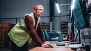 Un dipendente con indosso un giubbotto di sicurezza lavora concentrato davanti a uno schermo nel capannone, simbolo dell'ambiente di lavoro innovativo e tecnologico di KION Automation.