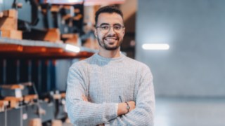 Un giovane collega con occhiali e orologio sta in piedi con sicurezza a braccia conserte nel magazzino ed è impegnato nel reparto messa in servizio di Linde. 