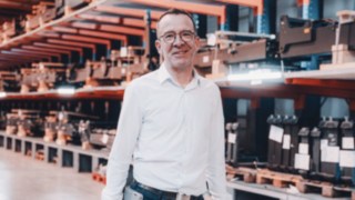 Un project manager in abiti da lavoro chiari si trova in un magazzino tecnico che rappresenta l'ambiente di lavoro in Linde nel settore dei progetti di automazione. 