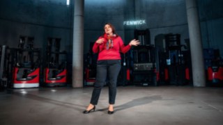 Una dipendente tiene una presentazione in un moderno ambiente industriale con carrelli elevatori Linde sullo sfondo, simbolo della collaborazione internazionale, dello scambio interculturale e delle opportunità di carriera globali in un'azienda interconnessa a livello mondiale.