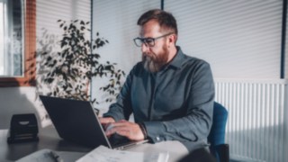 Un dipendente lavora concentrato al computer portatile in un ufficio luminoso con luce naturale, che simboleggia condizioni di lavoro flessibili che consentono un equilibrio tra vita privata e lavoro e un lavoro produttivo in home office o in ufficio.