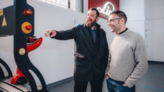 Due colleghi conversano davanti a un veicolo automatizzato Linde in un luminoso capannone industriale, simbolo di comunicazione aperta, lavoro di squadra tecnico e una cultura lavorativa caratterizzata dalla collaborazione e dal sostegno reciproco.