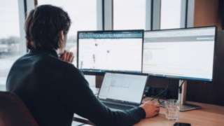 Un dipendente di KION Automation lavora su disegni tecnici e progetti di automazione a una scrivania con tre schermi, a simboleggiare l'uso delle tecnologie più moderne nell'intralogistica.