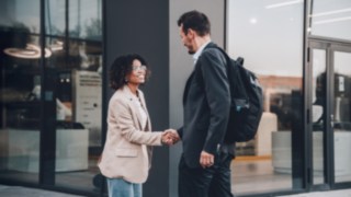 Due uomini d'affari si salutano con una stretta di mano davanti a un moderno edificio adibito a uffici, a simboleggiare la cooperazione internazionale, l'apertura culturale e le opportunità di carriera globali all'interno di un ambiente aziendale interconnesso.