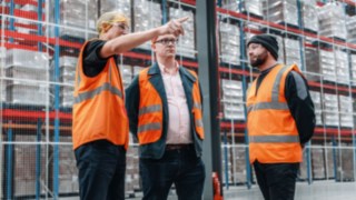 Tre dipendenti con giubbotti catarifrangenti conversano in un moderno magazzino a scaffali alti, incarnando la collaborazione orientata al lavoro di squadra, lo scambio professionale e una cultura aziendale collaborativa in un ambiente di lavoro dinamico.