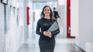 Una collaboratrice sicura di sé con un laptop in mano attraversa sorridente un moderno corridoio dell'ufficio e rappresenta il lavoro impegnato su progetti che rendono le catene di fornitura globali più efficienti e sostenibili.