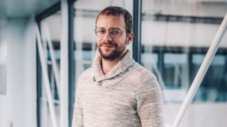 Un dipendente di KION Automation del reparto Solution Design sorride alla telecamera, ripreso davanti a un moderno edificio con facciata in vetro. 