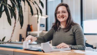 Una dipendente del reparto Solution Design di KION Automation sorride amichevolmente mentre lavora a una scrivania con un taglierino in un ufficio moderno. 