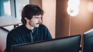 Un collega del reparto Solution Design di KION Automation lavora concentrato alla sua postazione di lavoro davanti a due monitor, circondato da una moderna lampada da terra e dalla luce del giorno.