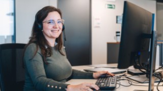 Una dipendente del reparto Solution Design di KION Automation sorride amichevolmente mentre lavora a una scrivania con un taglierino in un ufficio moderno. 