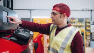 Un dipendente del reparto Commissioning di KION Automation, con giubbotto catarifrangente e cuffia rossa, è in piedi in modo professionale e cordiale in un magazzino a scaffali alti.