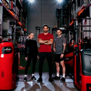 Una donna e due uomini in abbigliamento sportivo sono in piedi in un magazzino circondati da carrelli elevatori Linde Material Handling.